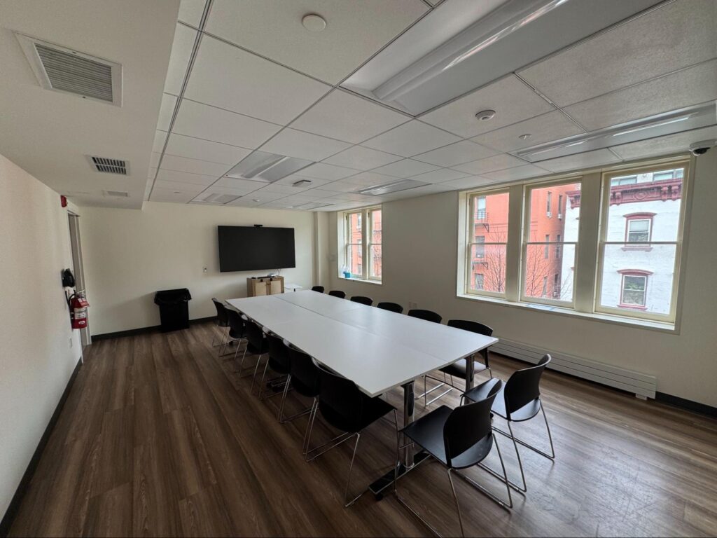 Middle Church Classroom set up boardroom style with a long white table, black chairs, and a wall-mounted screen beside tall windows