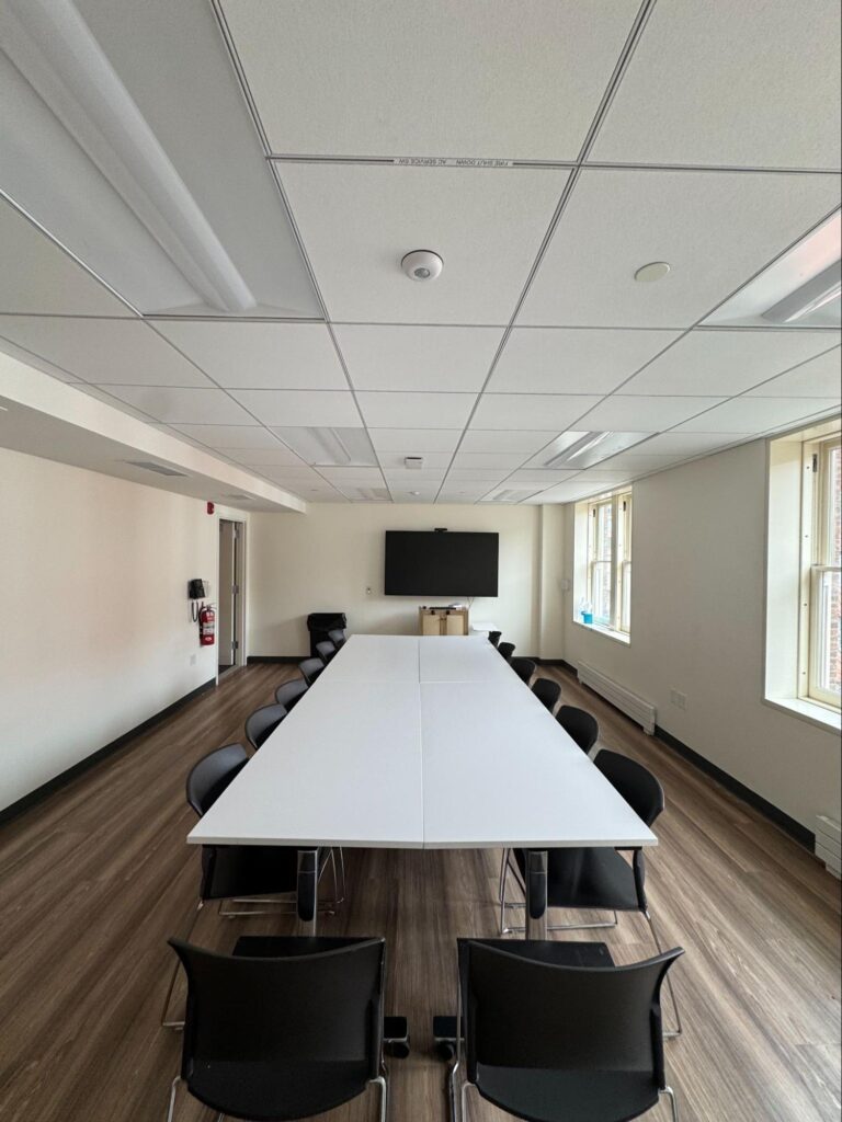 Middle Church Classroom boardroom table seen end-on, with a wall-mounted screen at the far end and daylight from the side windows