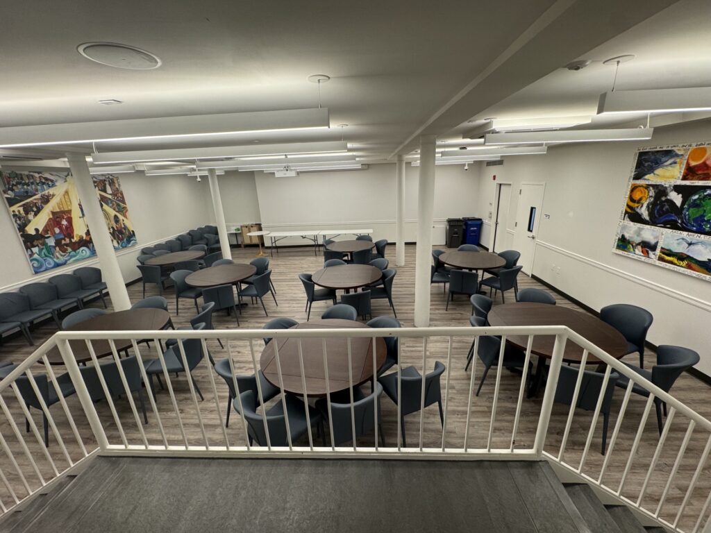 Middle Church Social Hall set with round banquet tables and upholstered chairs, viewed from the stairway, with community murals on the walls